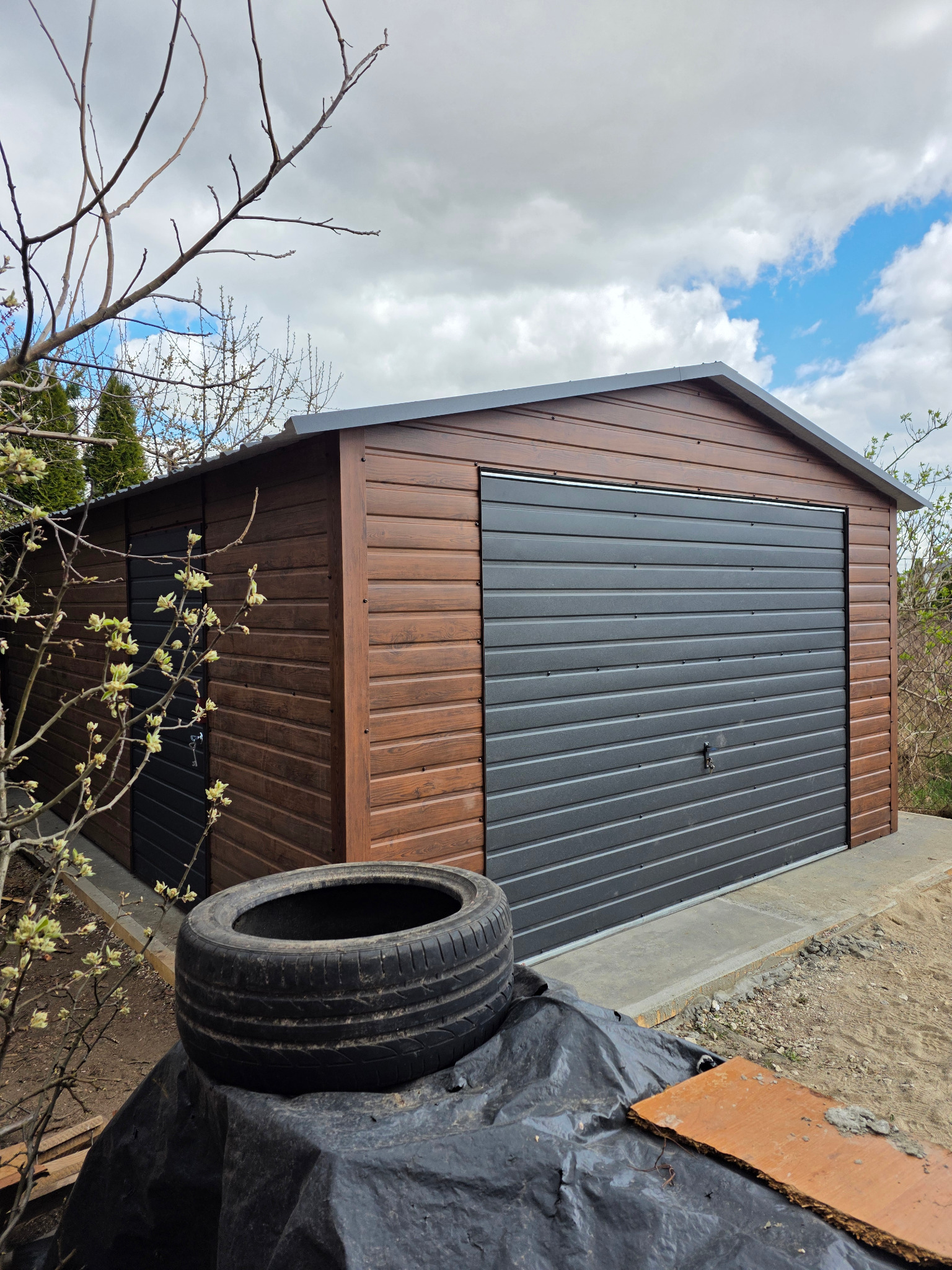 Drewnopodobny garaż blaszany z szarą bramą segmentową w Staroźrebach, widok z zewnątrz. Na pierwszym planie opona i folia budowlana.