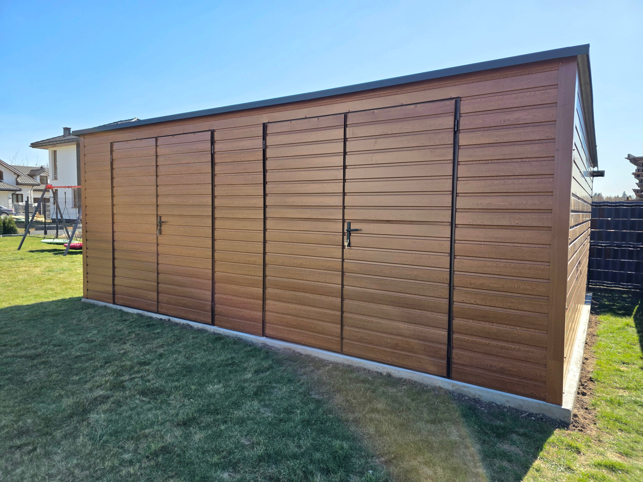 Brązowy garaż blaszany z trzema parami drzwi, imitujący drewno, na tle zielonego trawnika i błękitnego nieba. Widoczny fragment domu w tle.