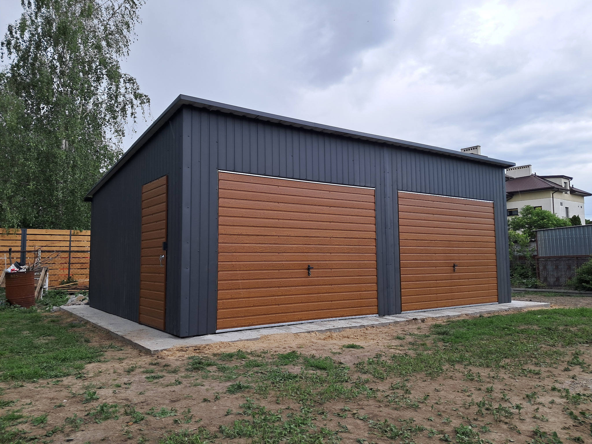 Dwustanowiskowy garaż blaszany z brązowymi bramami segmentowymi i szarą elewacją, na betonowej wylewce, widok z zewnątrz, pochmurne niebo w tle.
