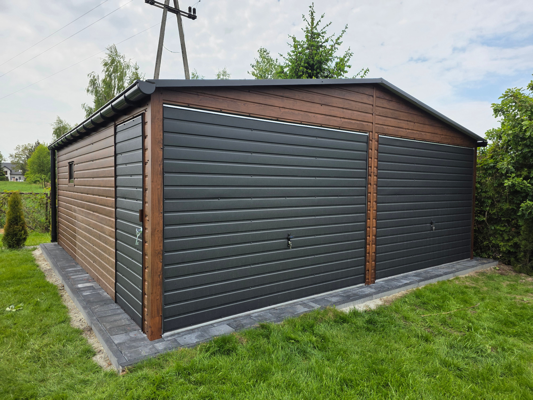Dwustanowiskowy garaż blaszany z brązową elewacją, czarnymi bramami segmentowymi i obrzeżem z kostki brukowej na trawniku. Widok z zewnątrz.