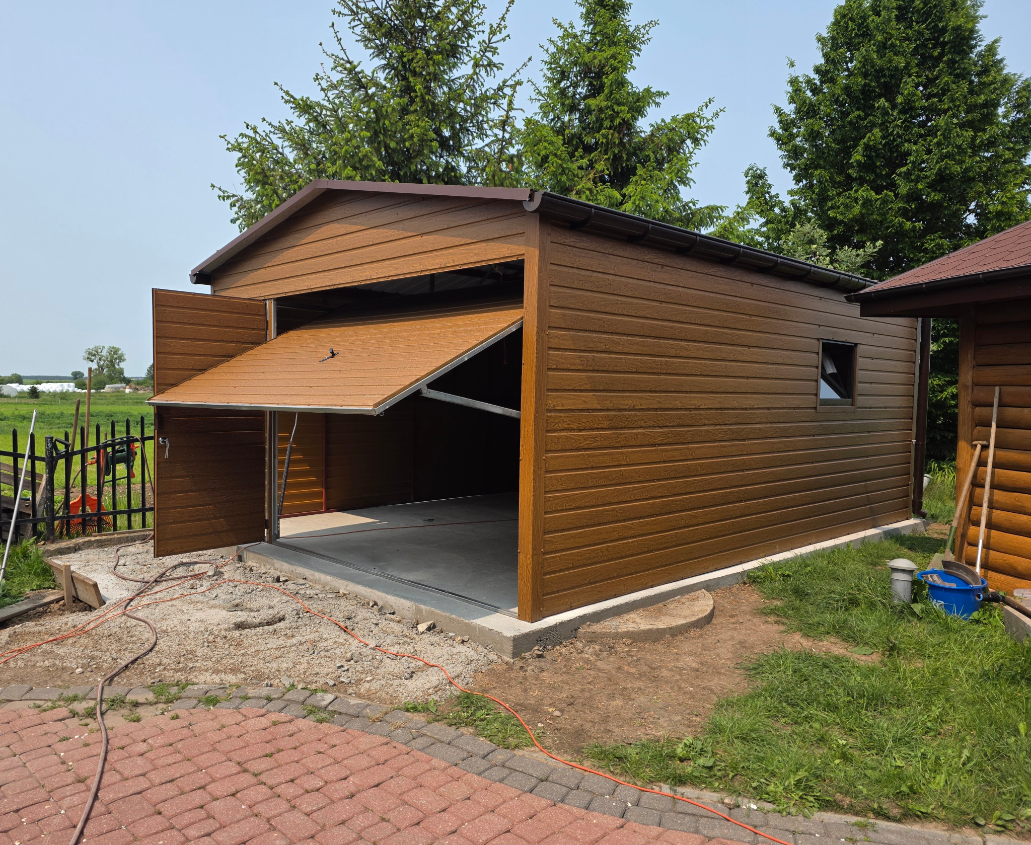 Brązowy garaż blaszany z otwartymi drzwiami segmentowymi i małym oknem, obok drewniana altana, kostka brukowa i zieleń w tle.