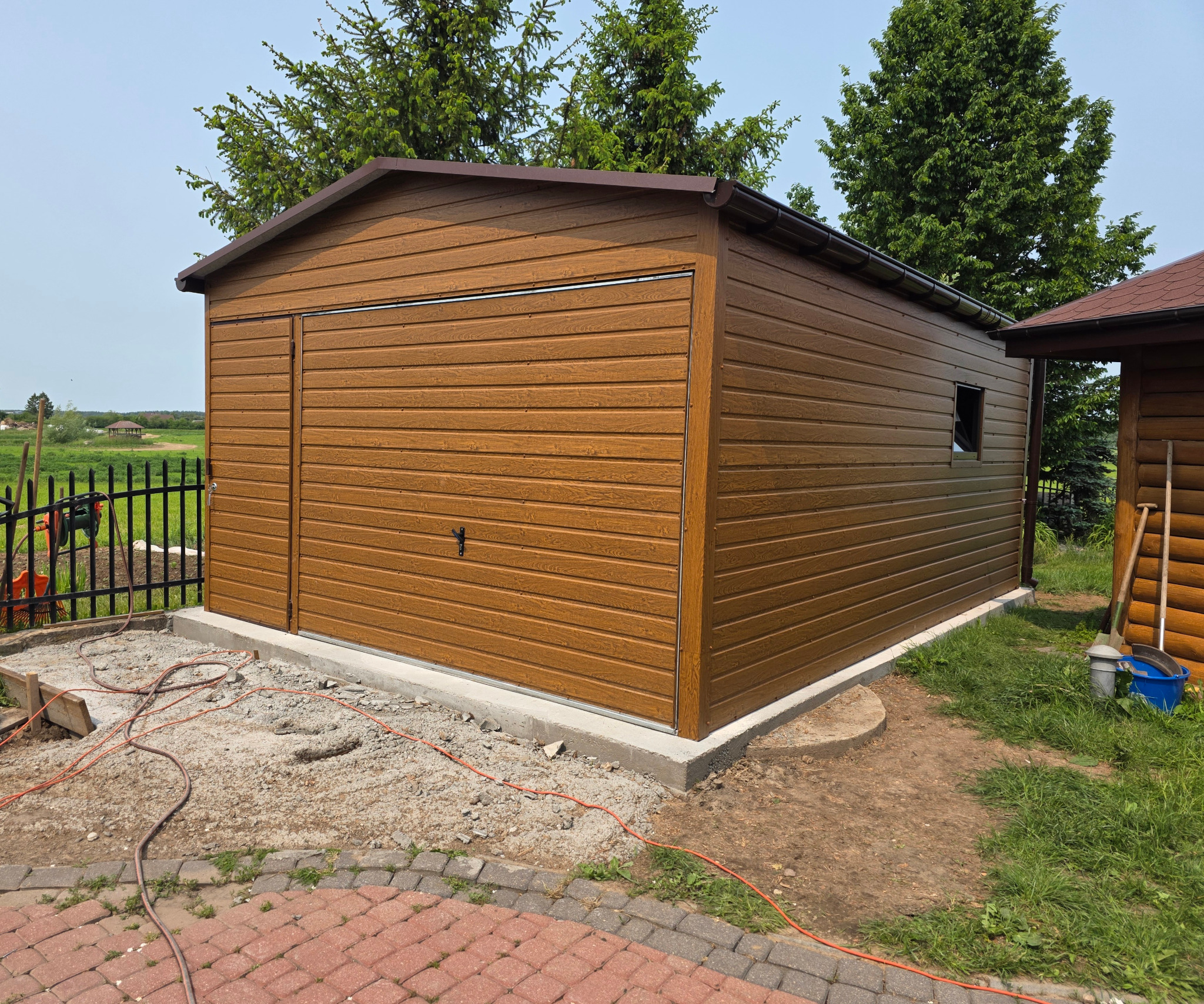 Brązowy garaż blaszany z imitacją drewna, widoczny z boku, z częściowo przygotowanym podłożem i kostką brukową w tle, w słoneczny dzień.