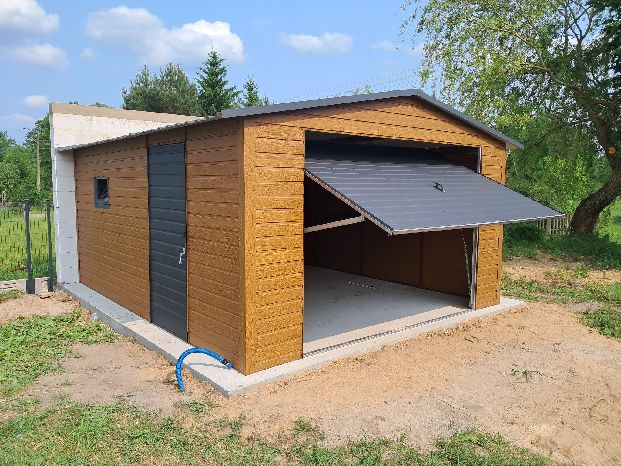 Drewnopodobny garaż blaszany z podnoszoną bramą, szarą stolarką i betonową podmurówką na tle zieleni i błękitnego nieba. Widoczny fragment ściany z bloczków.