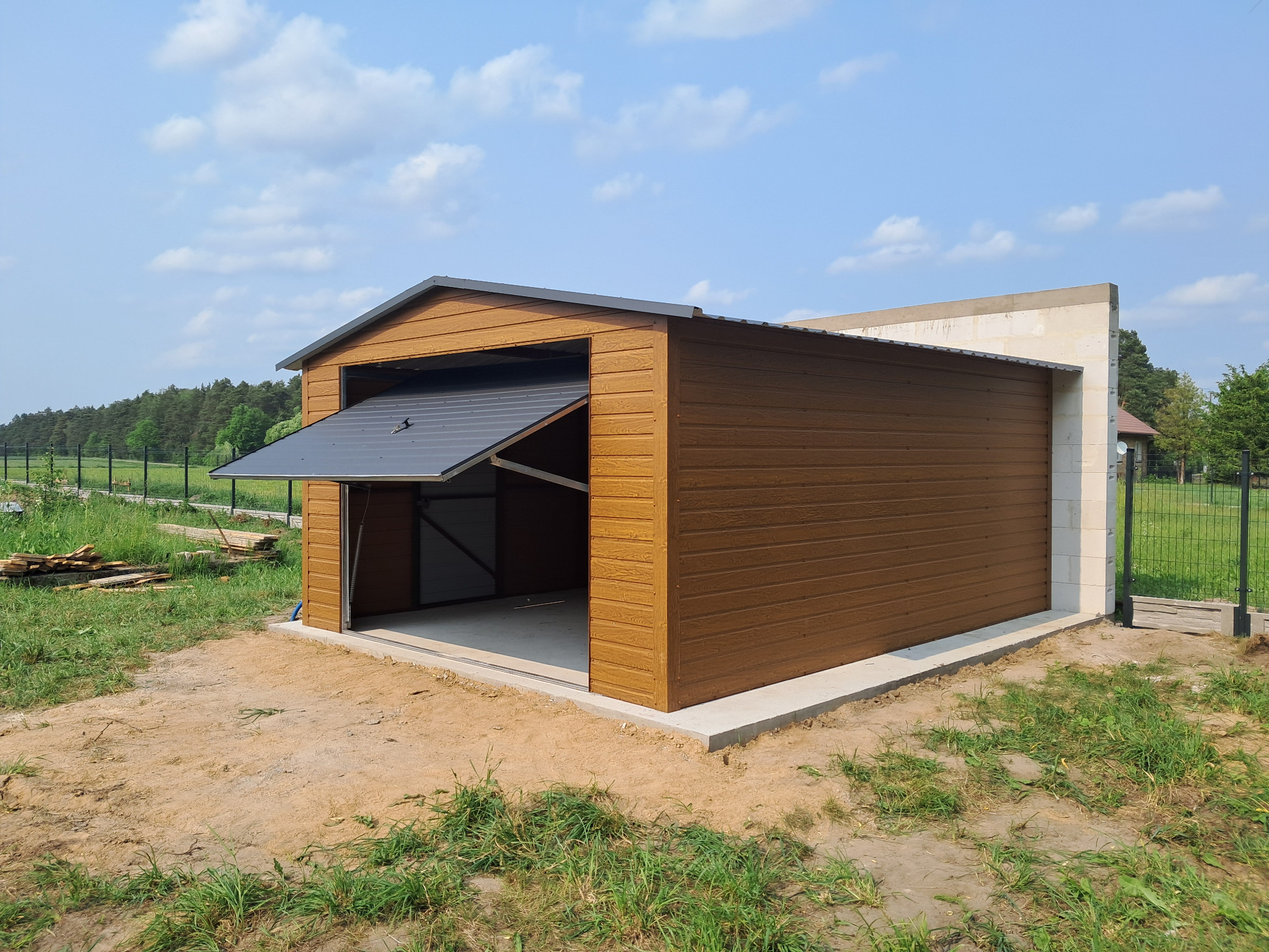 Otwarty garaż blaszany z brązowymi panelami, szarym dachem i betonową wylewką, na tle zieleni i błękitnego nieba z chmurami. Widoczne drewniane deski obok.