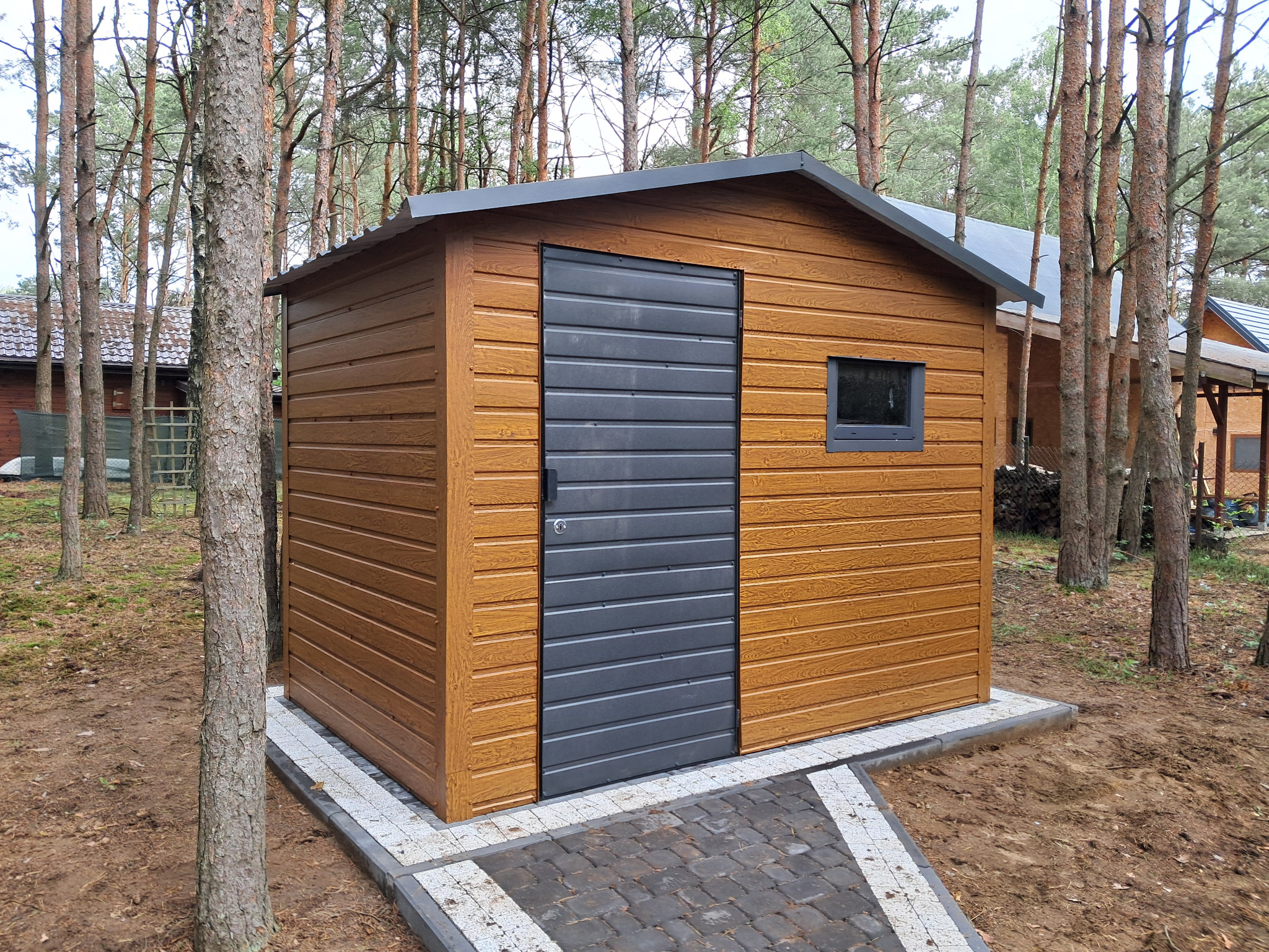 Drewniany domek narzędziowy z szarą, poziomą bramą, otoczony sosnowym lasem. Kostka brukowa ułożona jako ścieżka prowadzi do domku. Widoczny fragment dachu innego budynku.