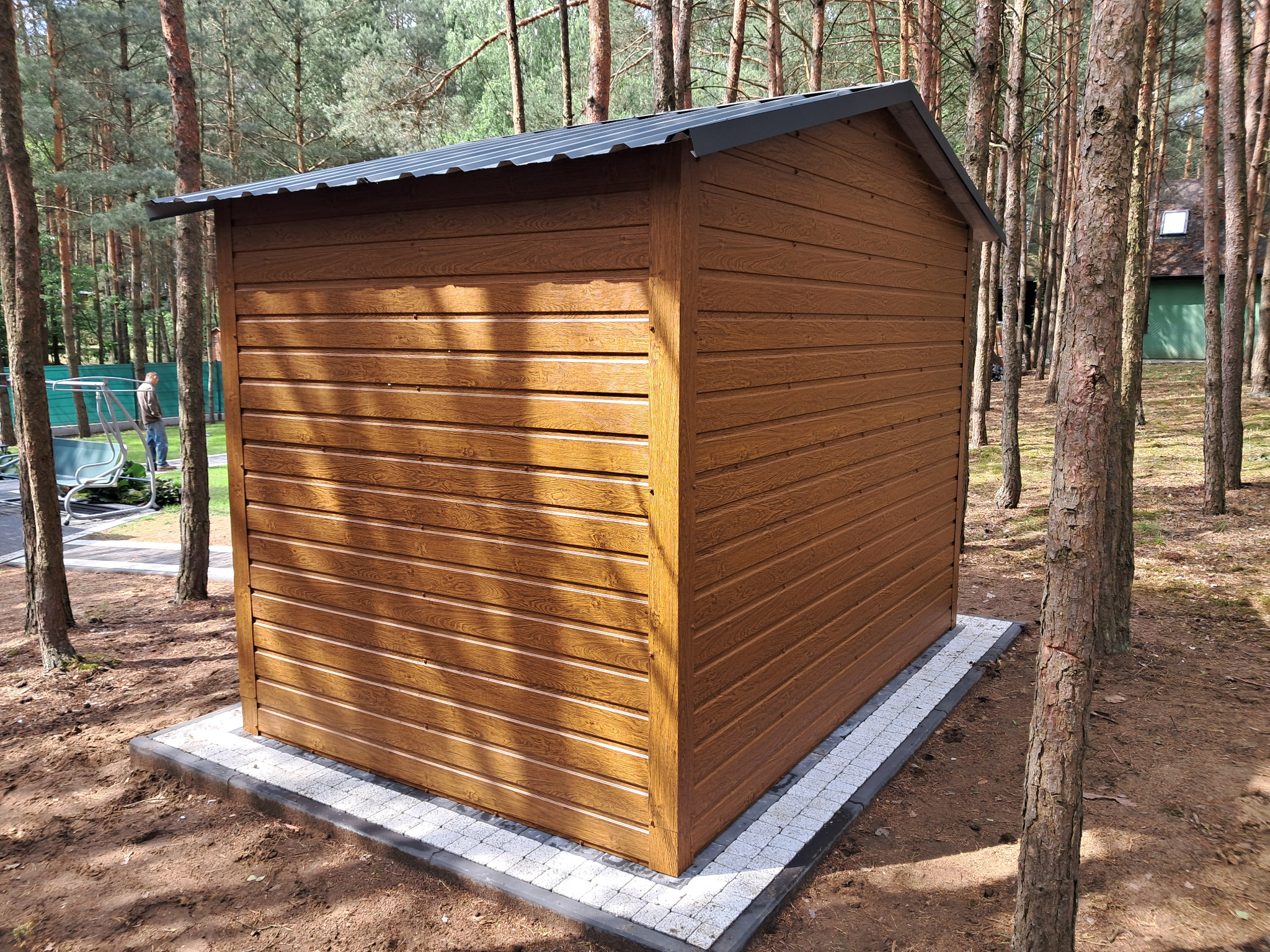 Brązowy, drewnopodobny garaż blaszany z szarą, spadzistą blachą, posadowiony na kostce brukowej w otoczeniu drzew iglastych. Widoczny fragment posesji w tle.