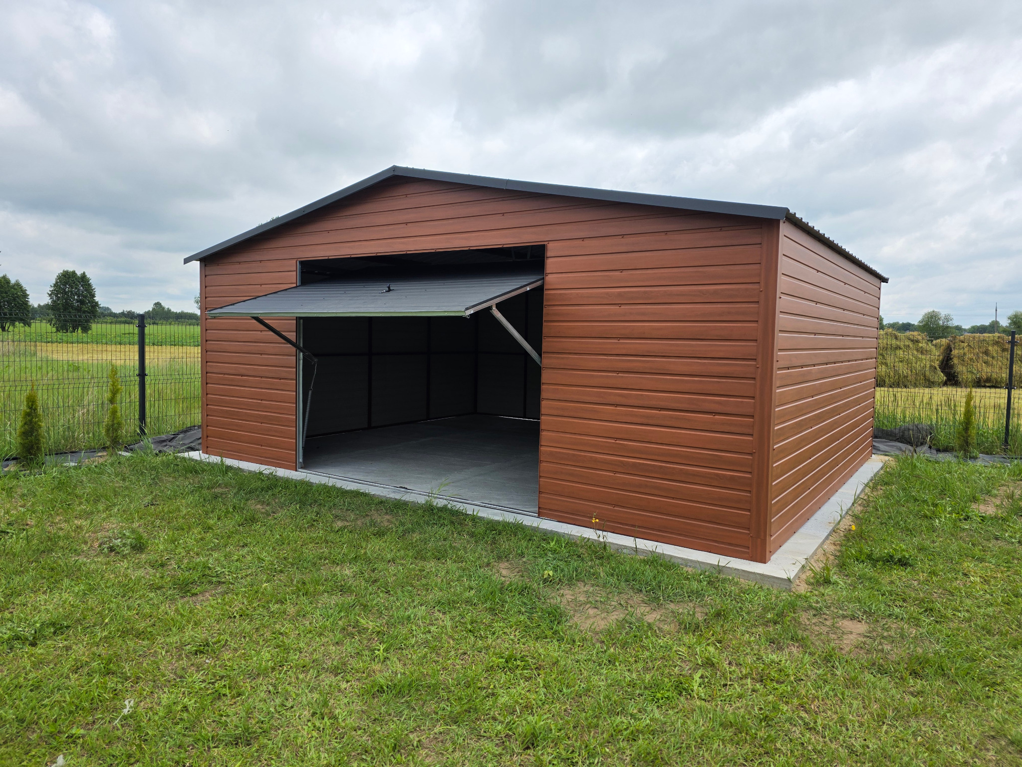 Brązowy garaż blaszany z otwartymi drzwiami uchylnymi, widoczny na tle zielonego trawnika i pochmurnego nieba. Konstrukcja z poziomych paneli, betonowa podstawa.