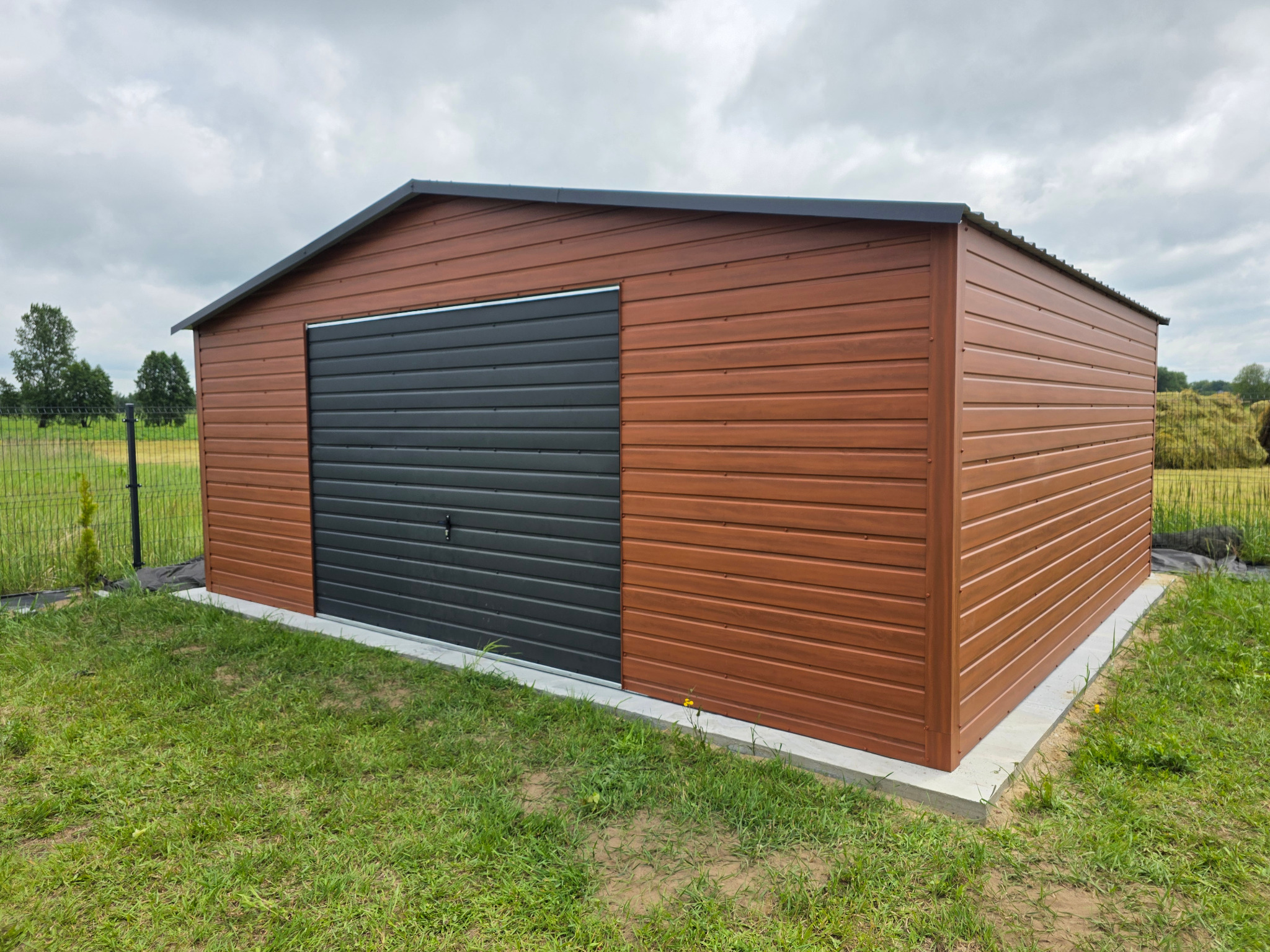 Brązowy garaż blaszany z szarą bramą segmentową na betonowej wylewce, otoczony zieloną trawą i ogrodzeniem z siatki w tle.