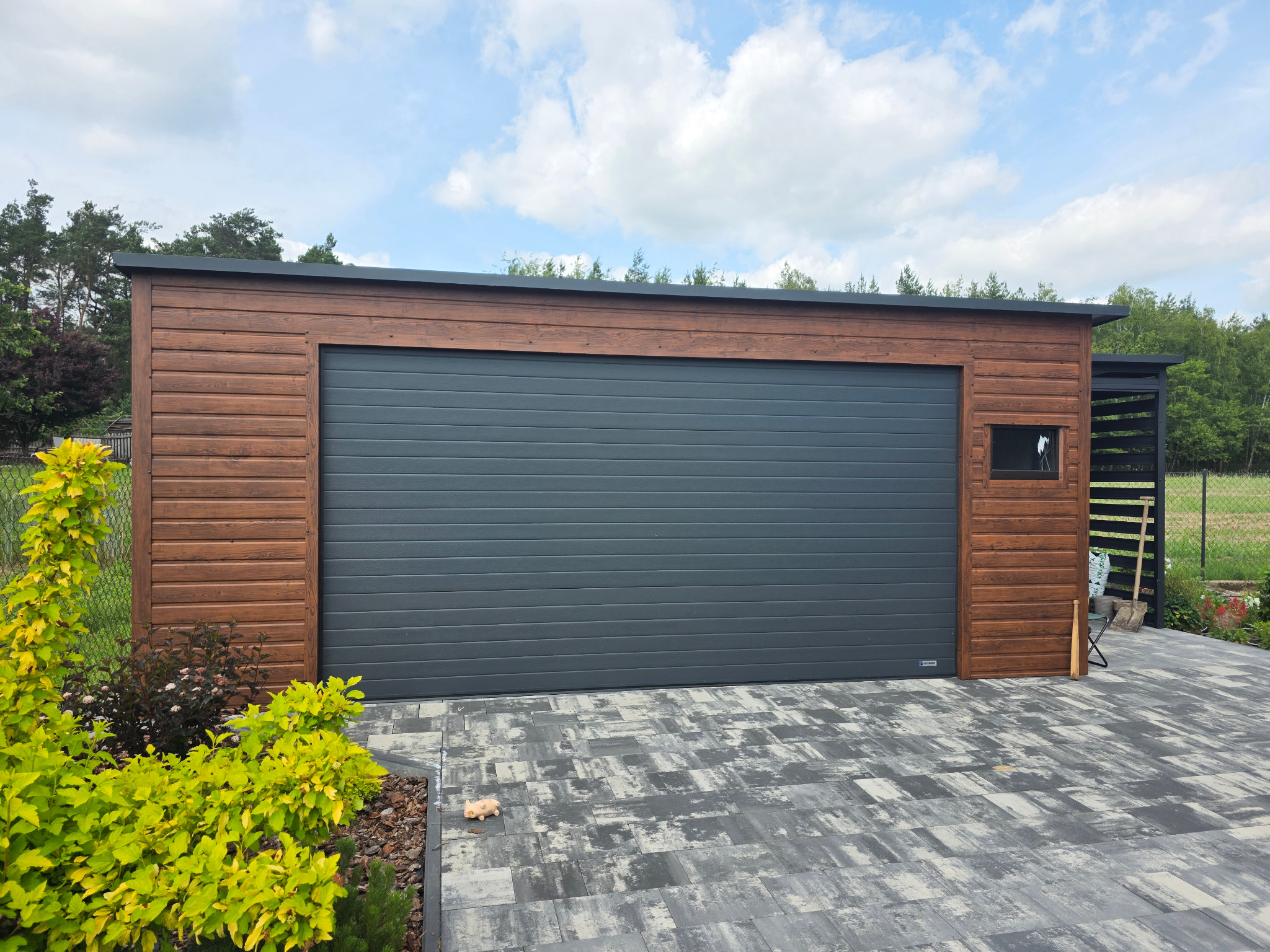 Nowoczesny garaż z drewnianą elewacją i szarą bramą segmentową, otoczony kostką brukową i zielenią. Widoczny fragment ogrodu w tle.