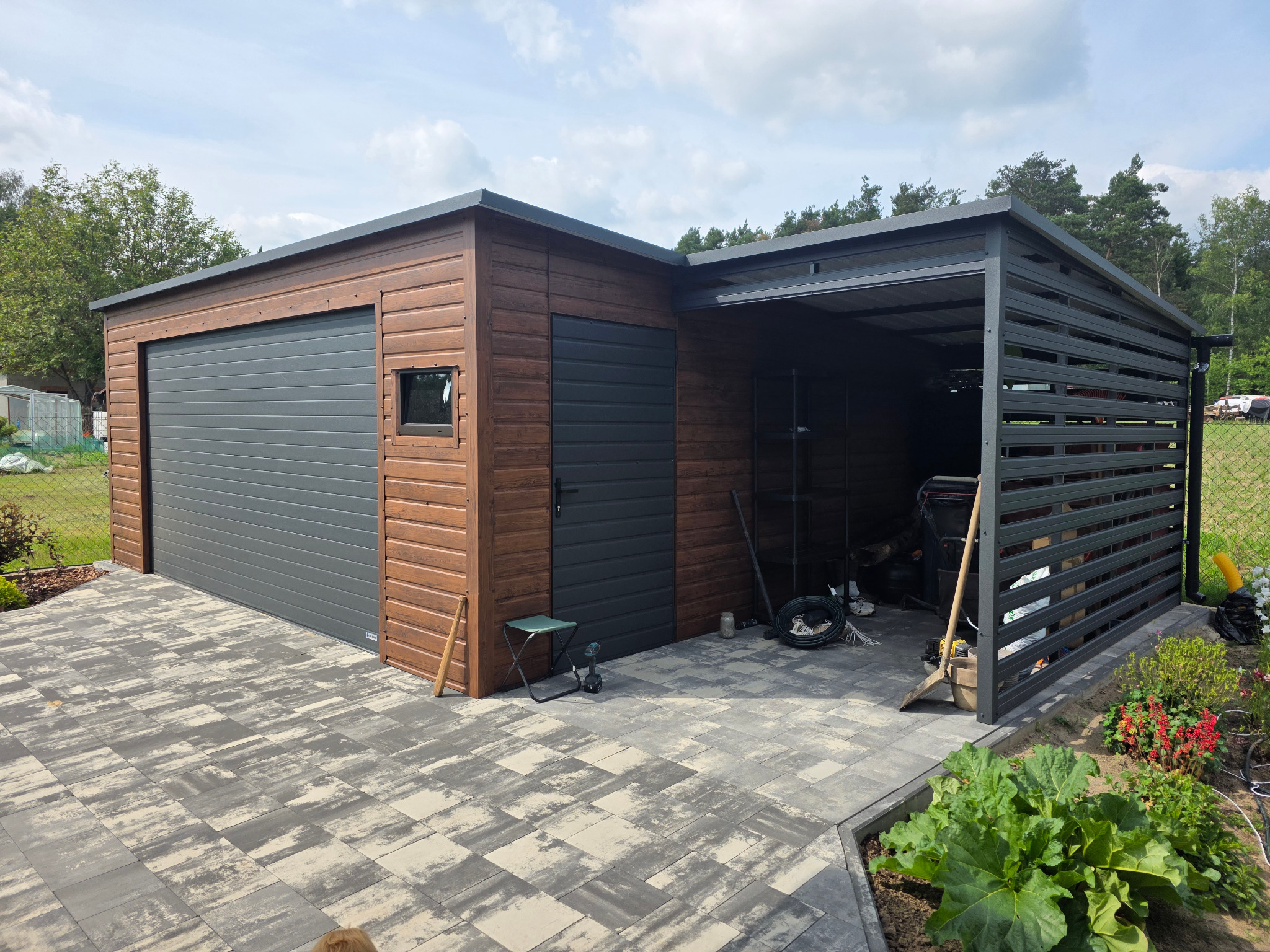 Nowoczesny garaż blaszany z bramą segmentową i otwartą wiatą, wykończony drewnopodobną elewacją, na posesji z kostką brukową i zielenią.