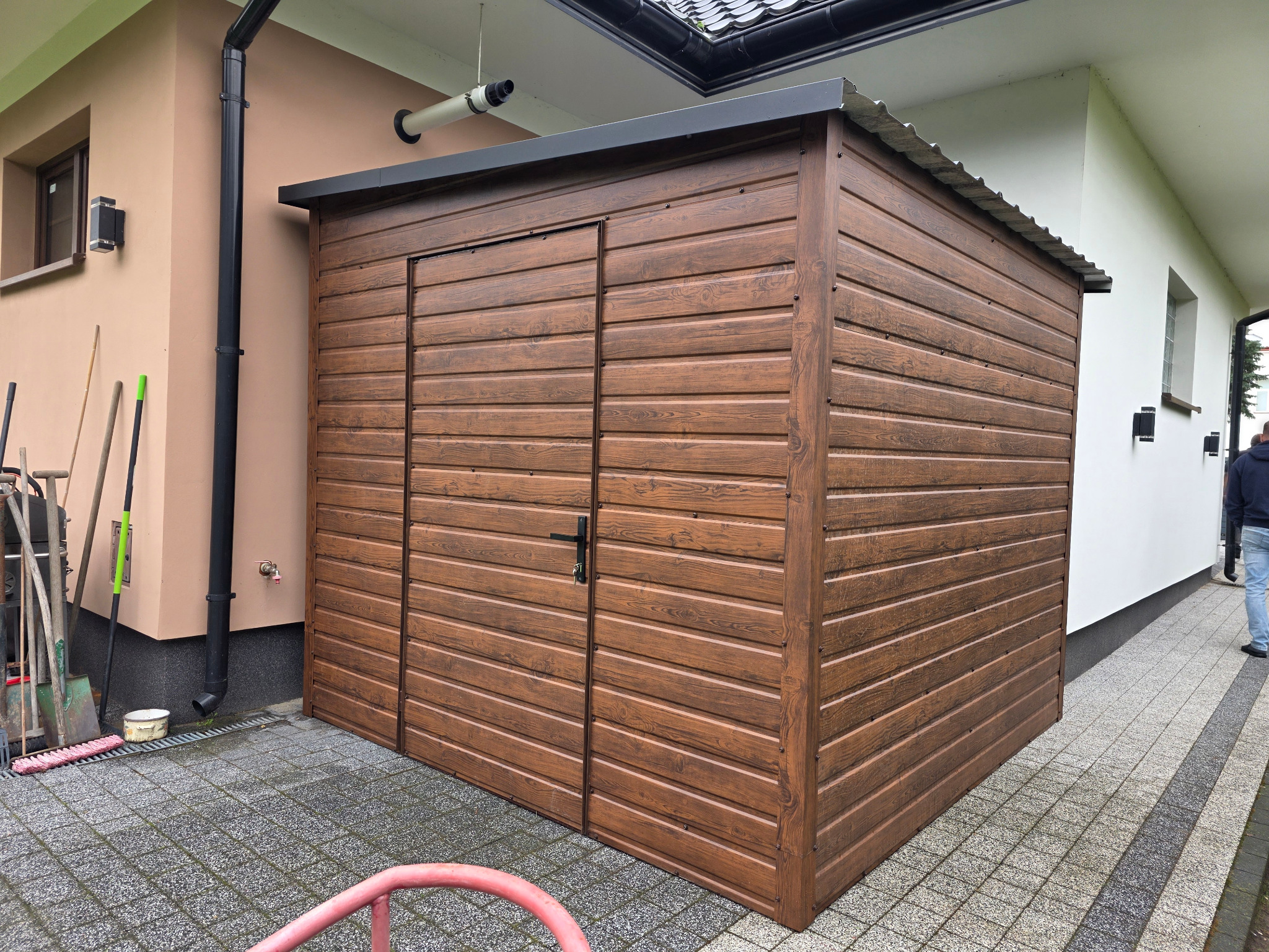 Brązowy, drewnopodobny garaż blaszany z dwuspadowym dachem, umieszczony na kostce brukowej, przylegający do budynku z widocznymi narzędziami ogrodowymi.