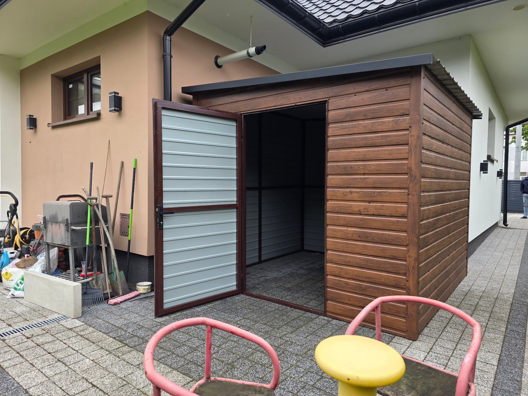 Drewniany domek narzędziowy z otwartymi drzwiami, umieszczony na kostce brukowej obok budynku. Widoczne narzędzia ogrodowe i grill. Uwagę przykuwa teleskop zamontowany pod dachem.