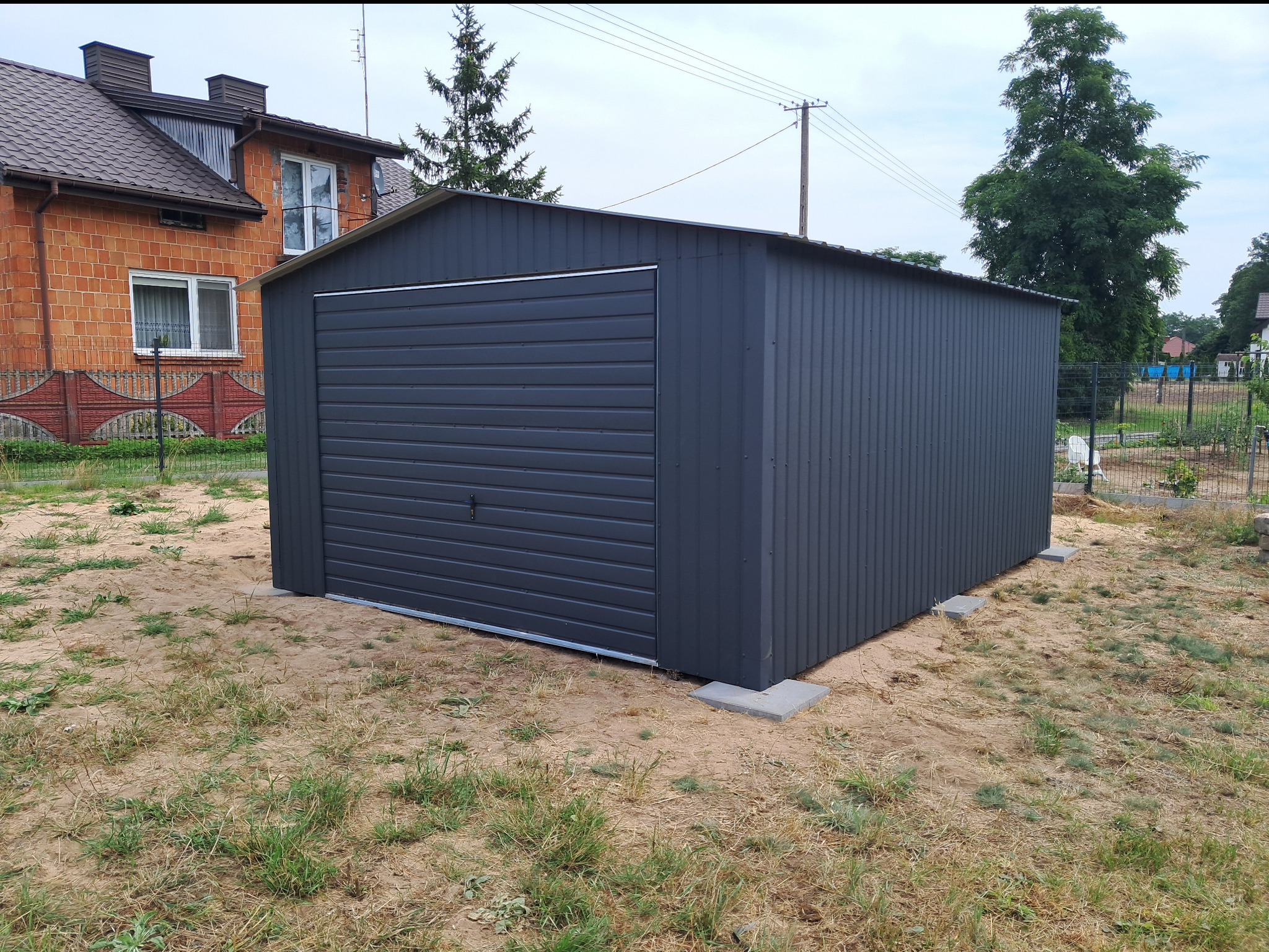 Nowoczesny, grafitowy garaż blaszany z bramą segmentową, ustawiony na betonowych płytach, na tle domu z czerwonej cegły i zieleni. Widok z przodu, w słoneczny dzień.