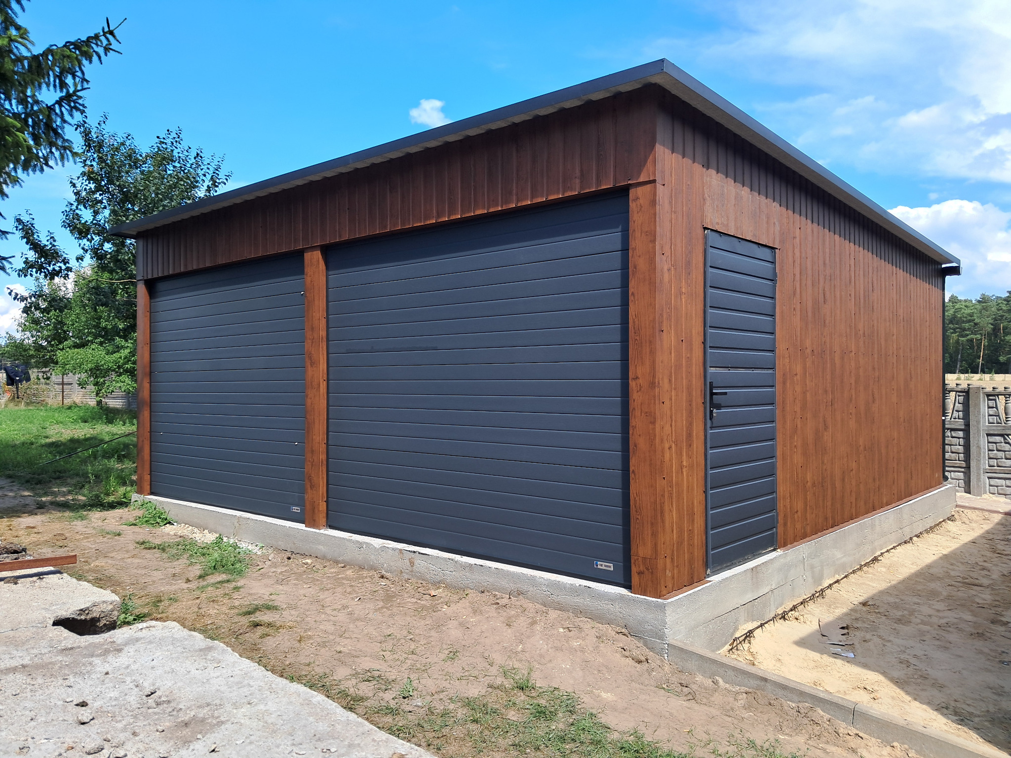 Nowoczesny garaż blaszany z dwoma bramami segmentowymi w kolorze grafitowym i drewnianą elewacją, z dodatkowym wejściem z boku, na betonowej podmurówce.