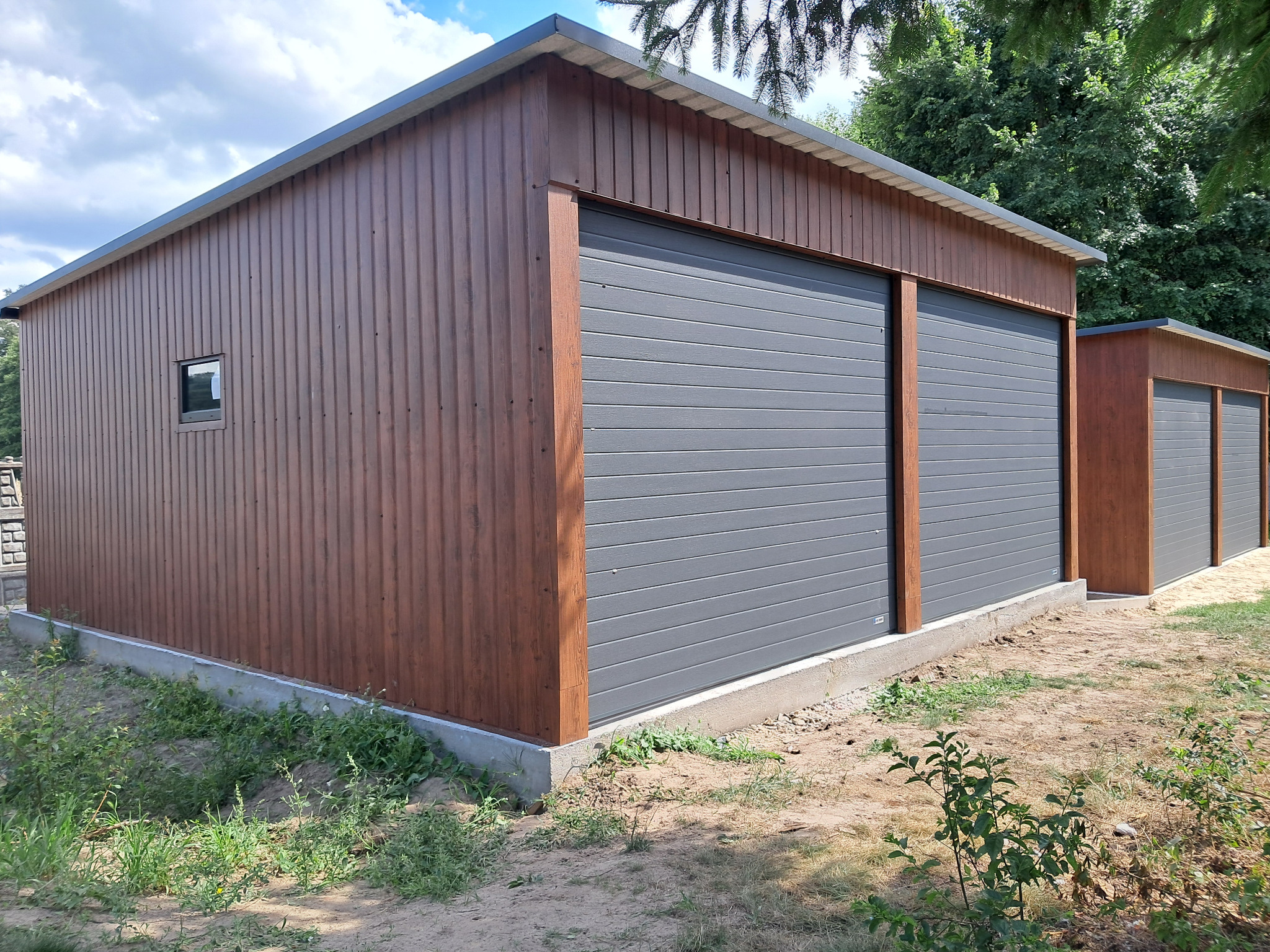 Dwa brązowe garaże blaszane z szarymi bramami segmentowymi, widoczne z zewnątrz na tle zieleni. Jeden garaż z oknem. Betonowa podmurówka.