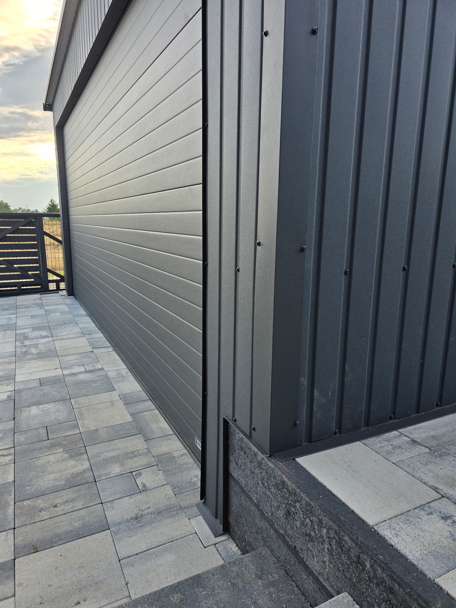 Szary garaż blaszany z poziomymi panelami, widok z boku, z kostką brukową w tle i fragmentem ogrodzenia. Detal wykończenia i połączenia paneli.