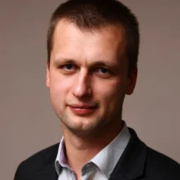 Portret mężczyzny w ciemnej marynarce i jasnej koszuli z wzorem, uśmiechającego się do kamery na neutralnym tle.