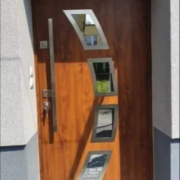 Brązowe drzwi wejściowe z czterema geometrycznymi przeszkleniami o srebrnych obramowaniach i długim, pionowym uchwytem, zamontowane w szarej ścianie budynku z kostki brukowej u podstawy.