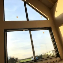 Wnętrze w stanie surowym z dużymi oknami o nietypowych kształtach (trójkąt i prostokąt), widok na krajobraz za oknem, ściany pokryte jasnym tynkiem.