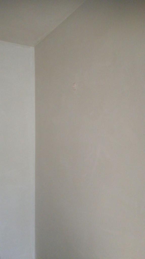Gładka, jasnoszara ściana po tynkowaniu maszynowym, widoczny fragment narożnika i mała nierówność na powierzchni.