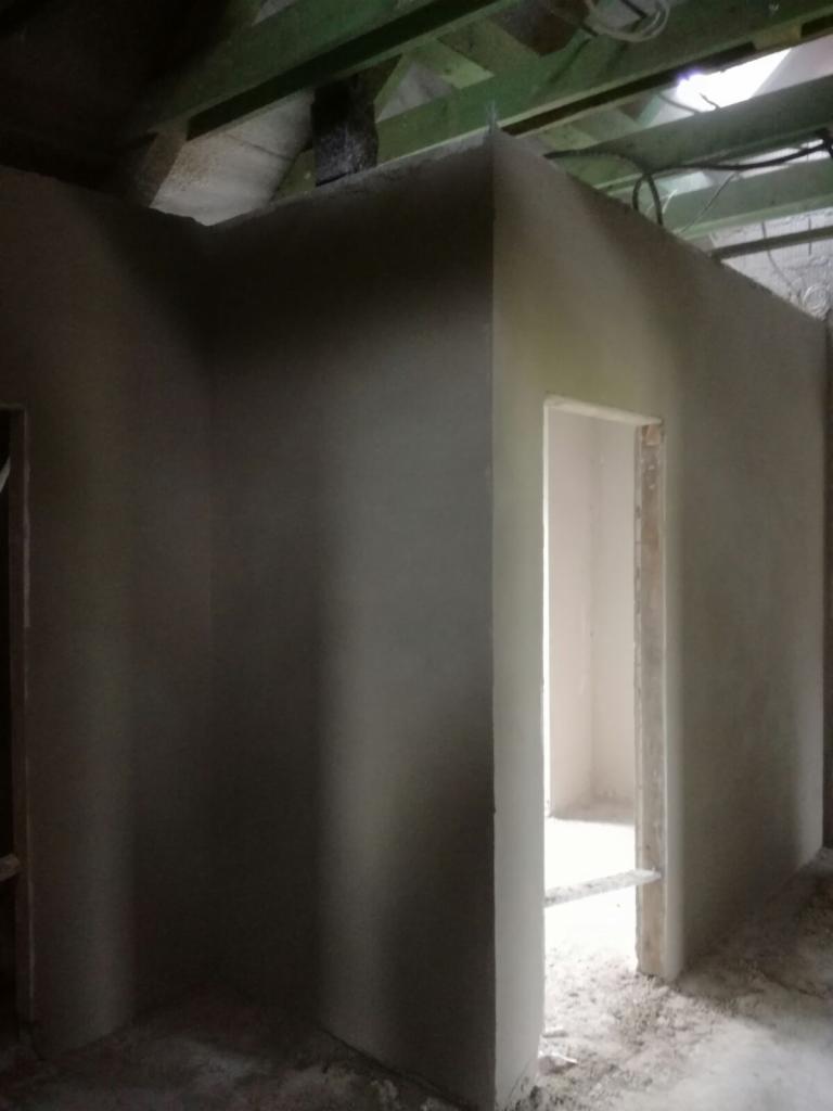 Wnętrze pomieszczenia w trakcie tynkowania maszynowego; ściany pokryte świeżym, szarym tynkiem, widoczne surowe drewniane elementy konstrukcyjne pod sufitem i niedokończone ościeże drzwiowe.
