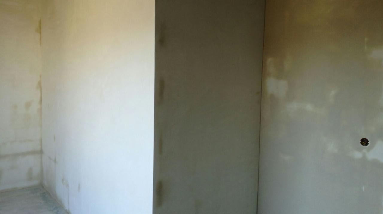 Świeżo nałożony tynk maszynowy na ścianach wewnętrznych pomieszczenia w trakcie schnięcia, widoczne nierówności i ślady po aplikacji, gniazdko elektryczne wystające ze ściany po prawej stronie.