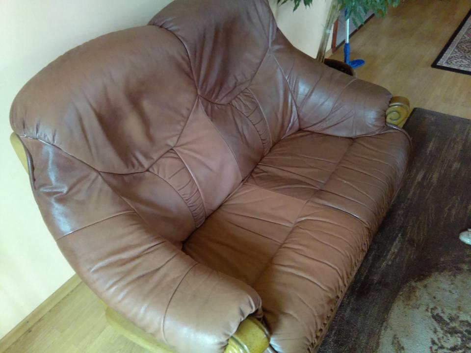 Przestarzały, brązowy, skórzany fotel z widocznymi śladami użytkowania i plamami na powierzchni, stojący na brudnej podłodze w pokoju z rośliną doniczkową w tle.