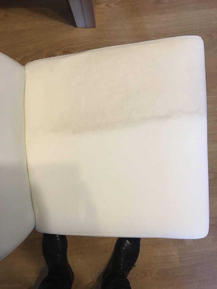 Białe krzesło tapicerowane z widocznym zabrudzeniem na siedzisku, stojące na drewnianej podłodze, z widocznymi butami osoby fotografującej.