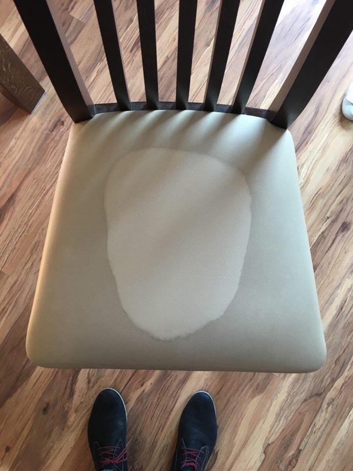 Krzesło z beżowym siedziskiem z wyraźną, okrągłą plamą po zalaniu, widoczne ciemne nogi krzesła i fragment podłogi z paneli oraz buty osoby fotografującej.