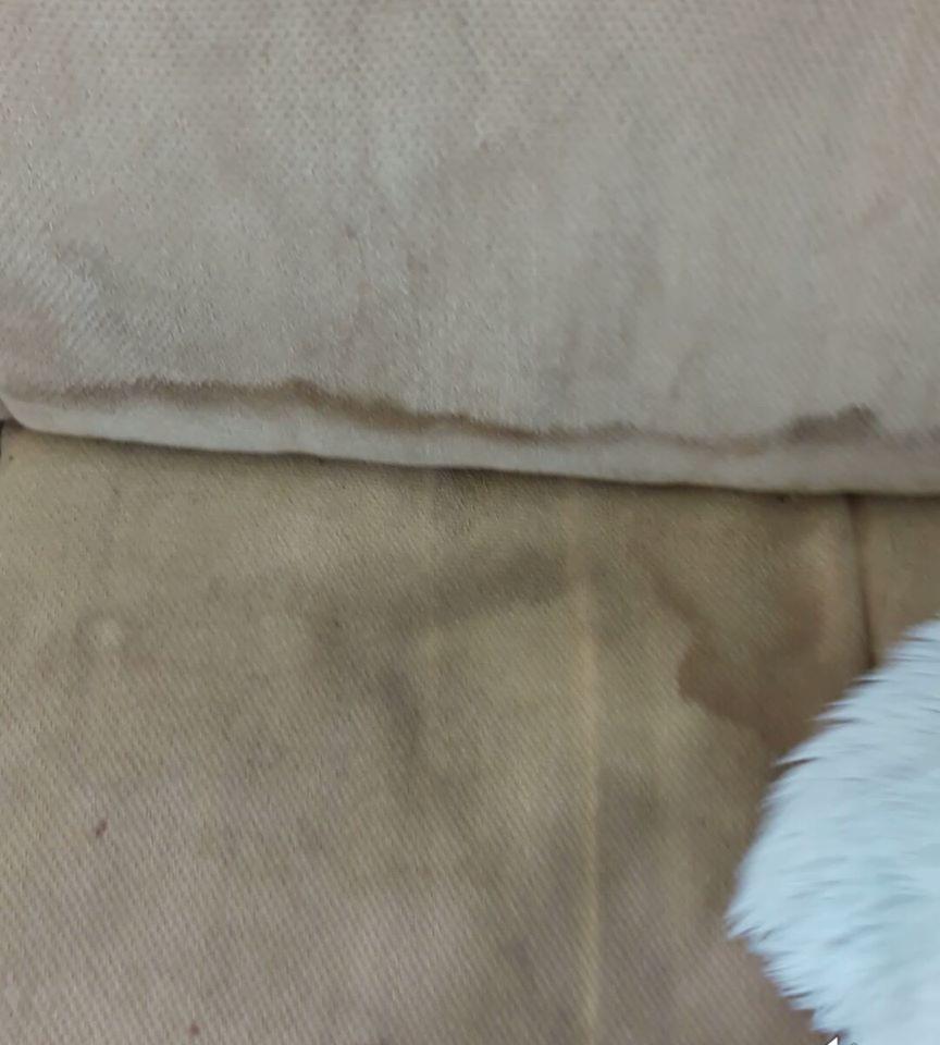 Fragment beżowej, tekstylnej sofy z widocznymi plamami i zabrudzeniami, częściowo przysłoniętej białym, puszystym materiałem.