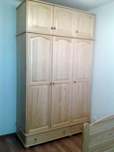 Wysoka, drewniana szafa trzydrzwiowa z nadstawką, z jasnego drewna, z małymi okrągłymi uchwytami, widoczna we Włocławku, z fragmentem ramy łóżka po prawej stronie.