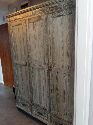 Trzydrzwiowa, drewniana szafa w stylu vintage z widocznym usłojeniem i delikatnymi zdobieniami, wykończona w odcieniach szarości i brązu, widziana z perspektywy wnętrza.