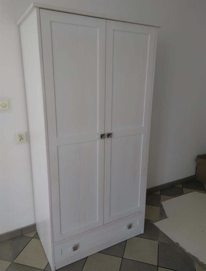 Dwudrzwiowa, biała szafa z dwiema szufladami na dole, z prostymi, kwadratowymi uchwytami, na tle białej ściany i podłogi z szaro-beżowych płytek.