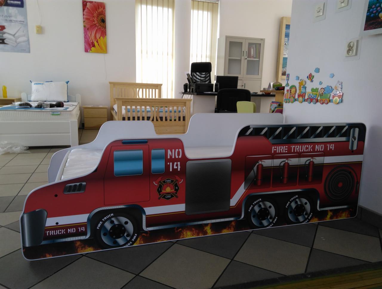 Łóżko dziecięce w kształcie wozu strażackiego z nadrukiem 'Fire Truck No 14', stojące w jasnym pomieszczeniu ekspozycyjnym z innymi łóżkami i biurkiem w tle.