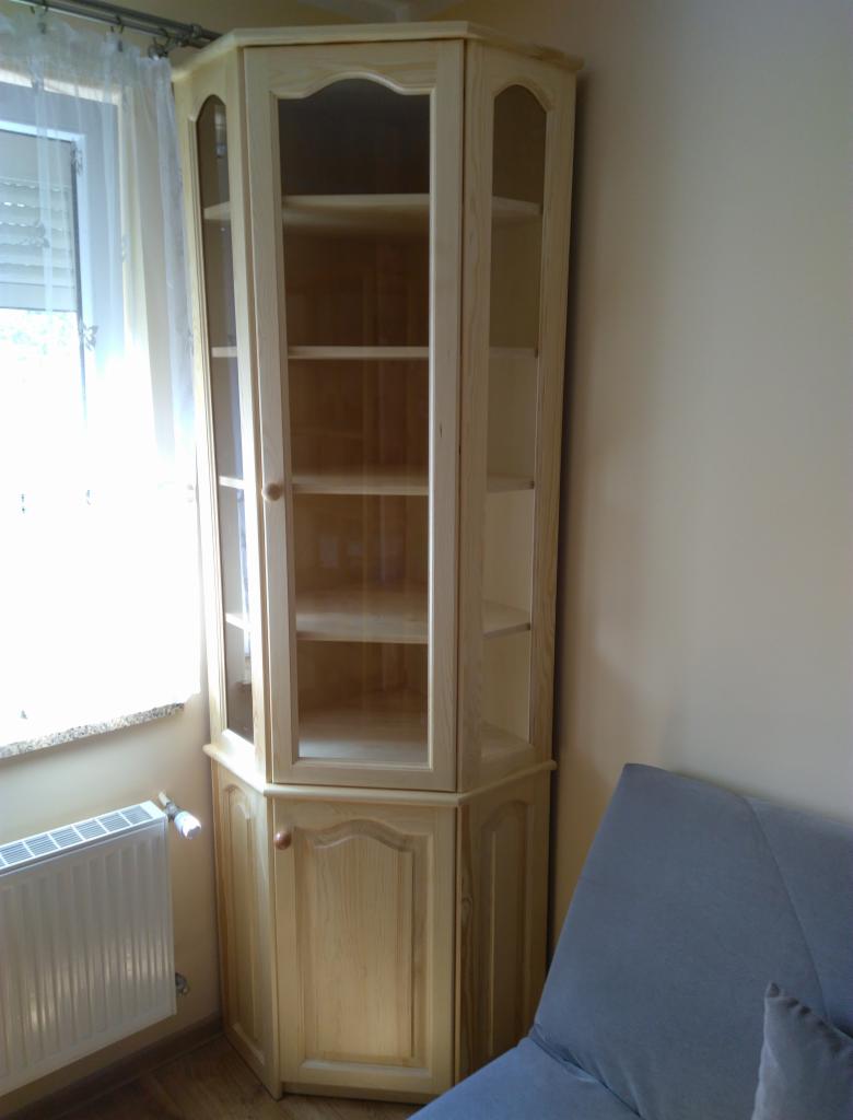 Narożna, drewniana witryna z przeszklonymi drzwiami, usytuowana w rogu pokoju obok okna i grzejnika.