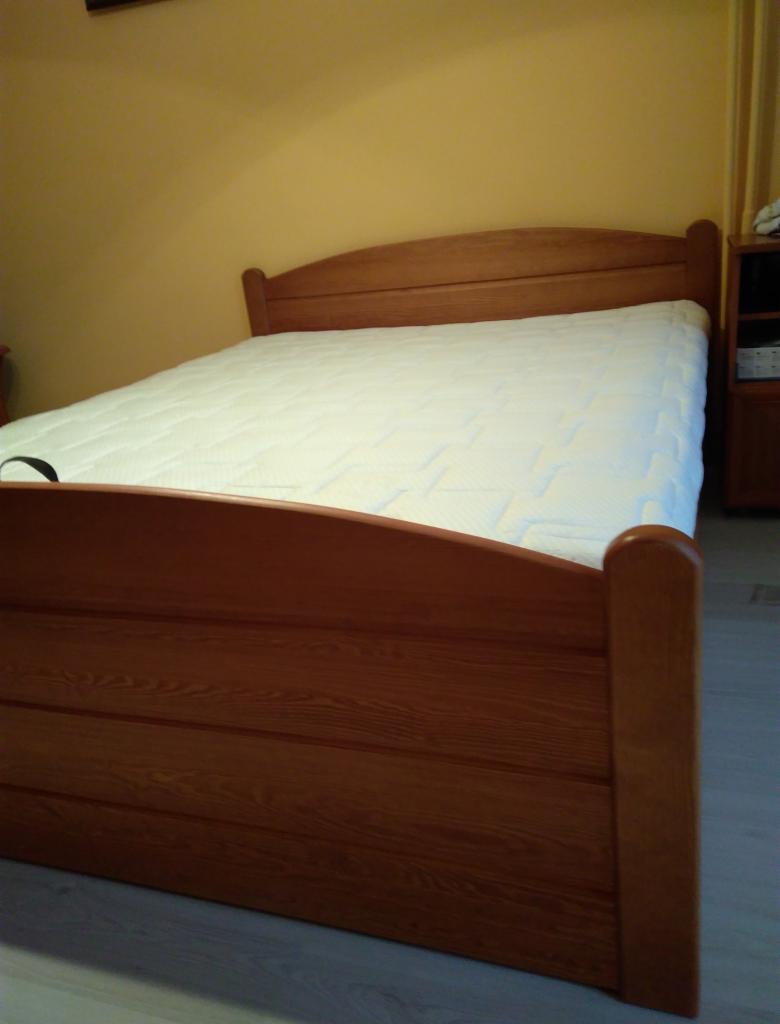 Drewniane łóżko z materacem w pokoju o żółtych ścianach, widoczne szafki nocne w tle.