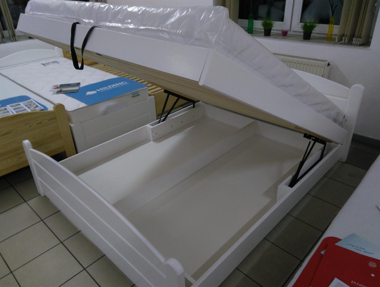 Białe łóżko z podnoszonym stelażem i pojemnikiem na pościel, widoczne mechanizmy podnoszące, materac w folii ochronnej, w tle inne łóżko i okno.