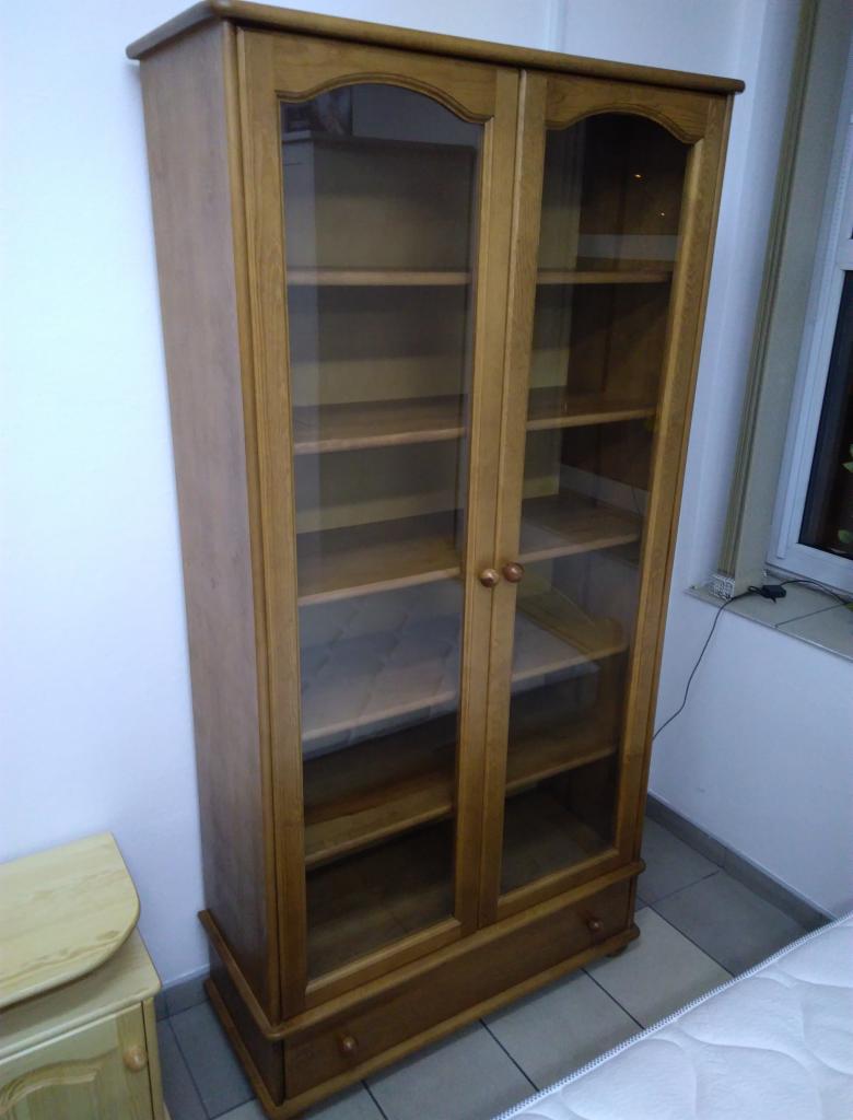 Drewniana witryna z przeszklonymi drzwiami, widoczne półki, dolna szuflada, styl klasyczny, jasne drewno, ustawiona w rogu pokoju obok okna i szafki nocnej.