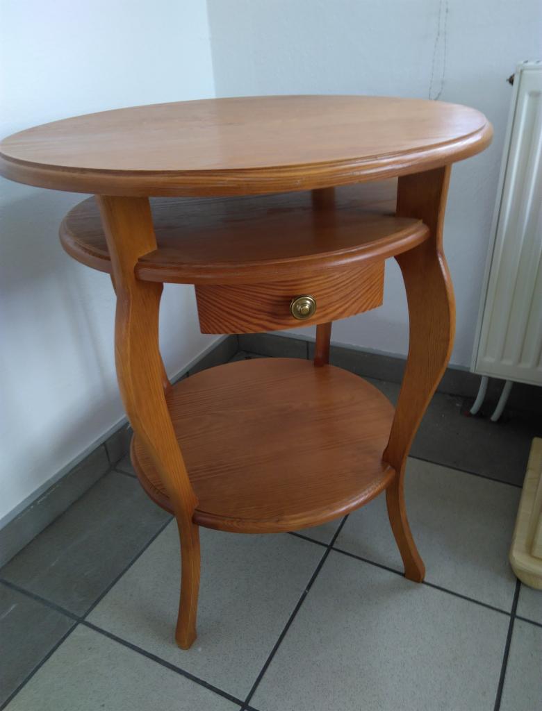 Okrągły, drewniany stolik pomocniczy z szufladą i dwiema półkami, widoczny na tle ściany i grzejnika.