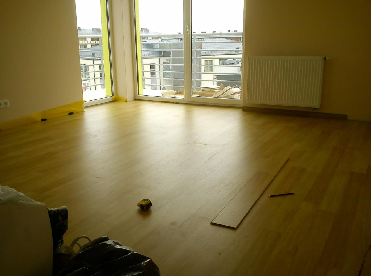 Jasne panele podłogowe w trakcie montażu, widoczna listwa przypodłogowa, okno balkonowe z widokiem na budynki i grzejnik na ścianie, taśma ochronna na ścianach, miarka i ołówek na podłodze.
