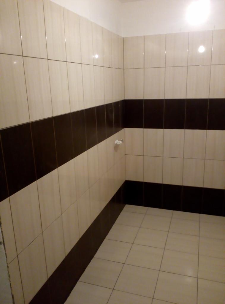 Wykończona łazienka z beżowo-brązowymi płytkami ułożonymi w poziome pasy, widoczny fragment ściany z uchwytem na papier toaletowy.