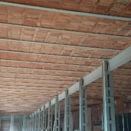 Wnętrze budynku w trakcie renowacji: ceglany sufit z metalowymi belkami nośnymi, widoczne ślady po starych tynkach na cegłach.