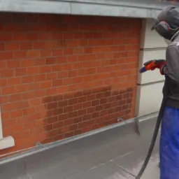 Pracownik w stroju ochronnym piaskuje czerwoną ceglaną ścianę budynku z białymi obramowaniami okien, widoczne efekty czyszczenia na powierzchni cegieł.