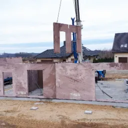 Montaż ścian domu z prefabrykatów keramzytowych za pomocą dźwigu, widoczne logo firmy na ścianach, w tle zabudowa jednorodzinna.