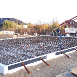 Przygotowanie płyty fundamentowej zbrojonej siatką stalową i izolacją termiczną, widoczna pompa do betonu w tle i rury kanalizacyjne wystające z płyty.