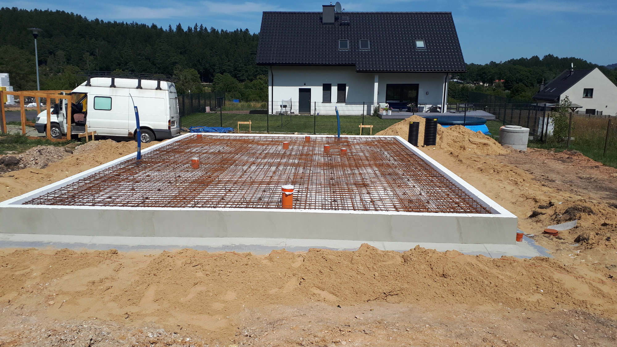 Przygotowany fundament z siatką zbrojeniową i pionowymi rurami instalacyjnymi, obok zaparkowany biały bus z bagażnikiem dachowym, w tle dom jednorodzinny z ciemnym dachem i zielenią.