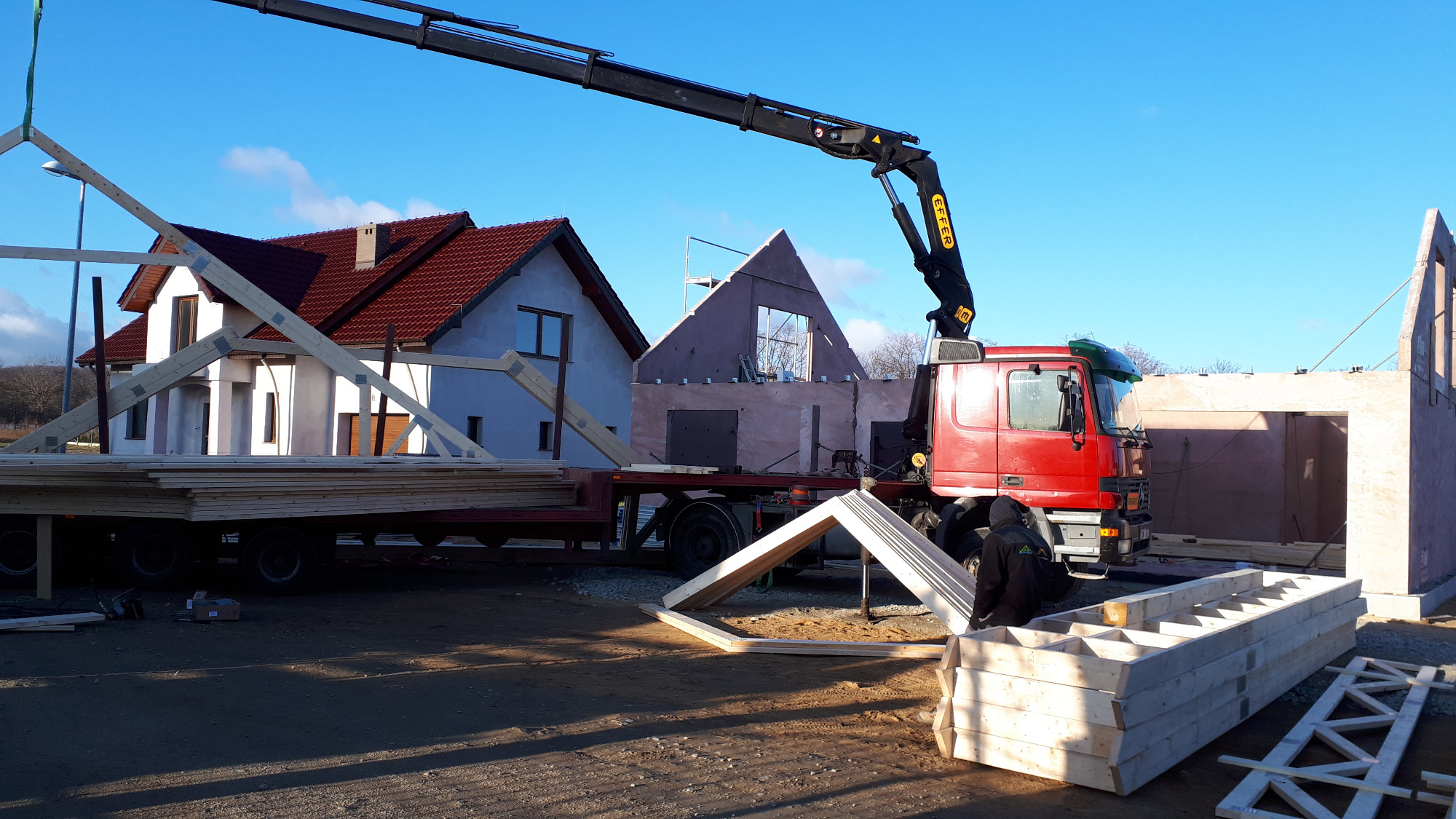 Montaż drewnianej konstrukcji domu z użyciem dźwigu samochodowego na tle istniejącego budynku i domu w trakcie budowy.