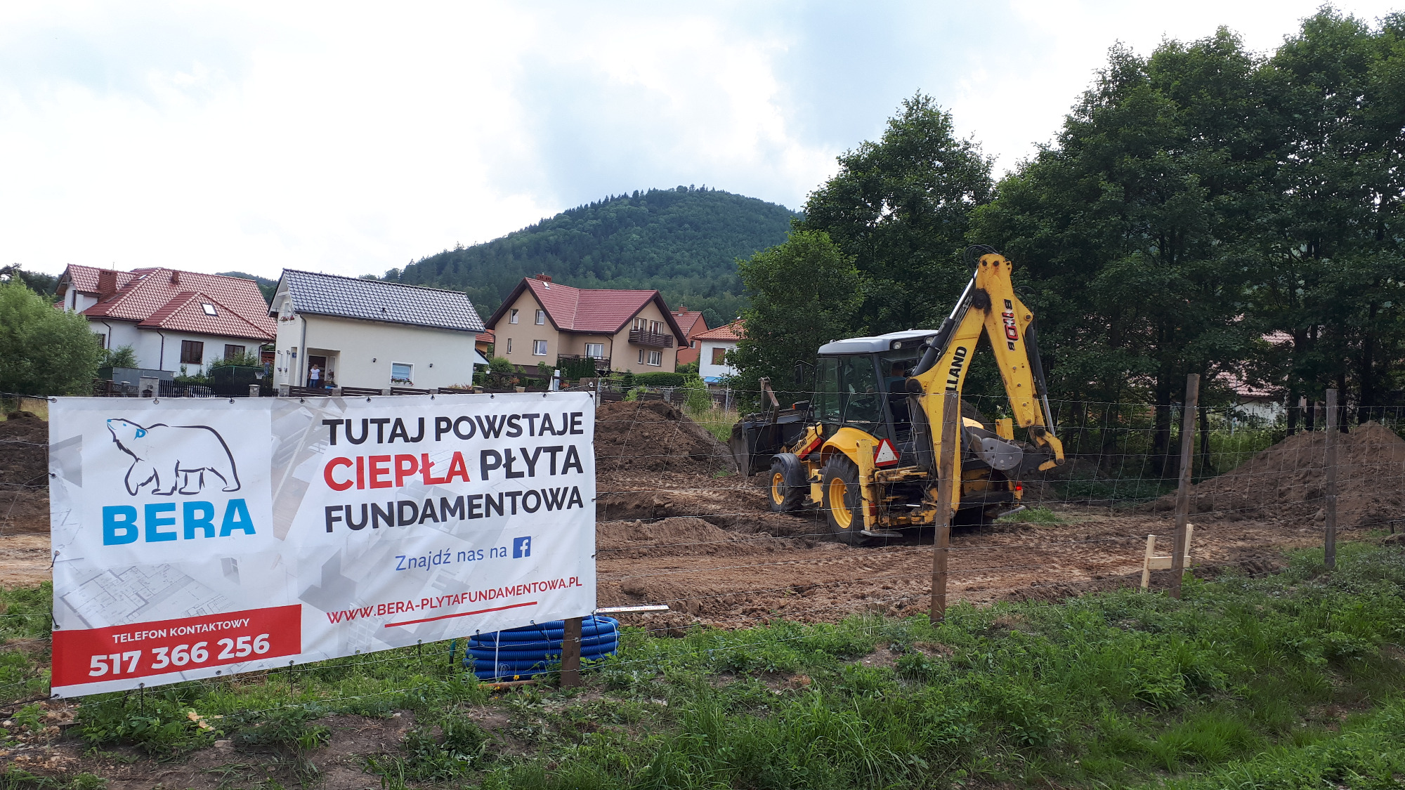 Żółta koparko-ładowarka na placu budowy, w tle wzniesienia i zabudowa mieszkalna, na pierwszym planie baner reklamowy firmy budowlanej oferującej ciepłe płyty fundamentowe.