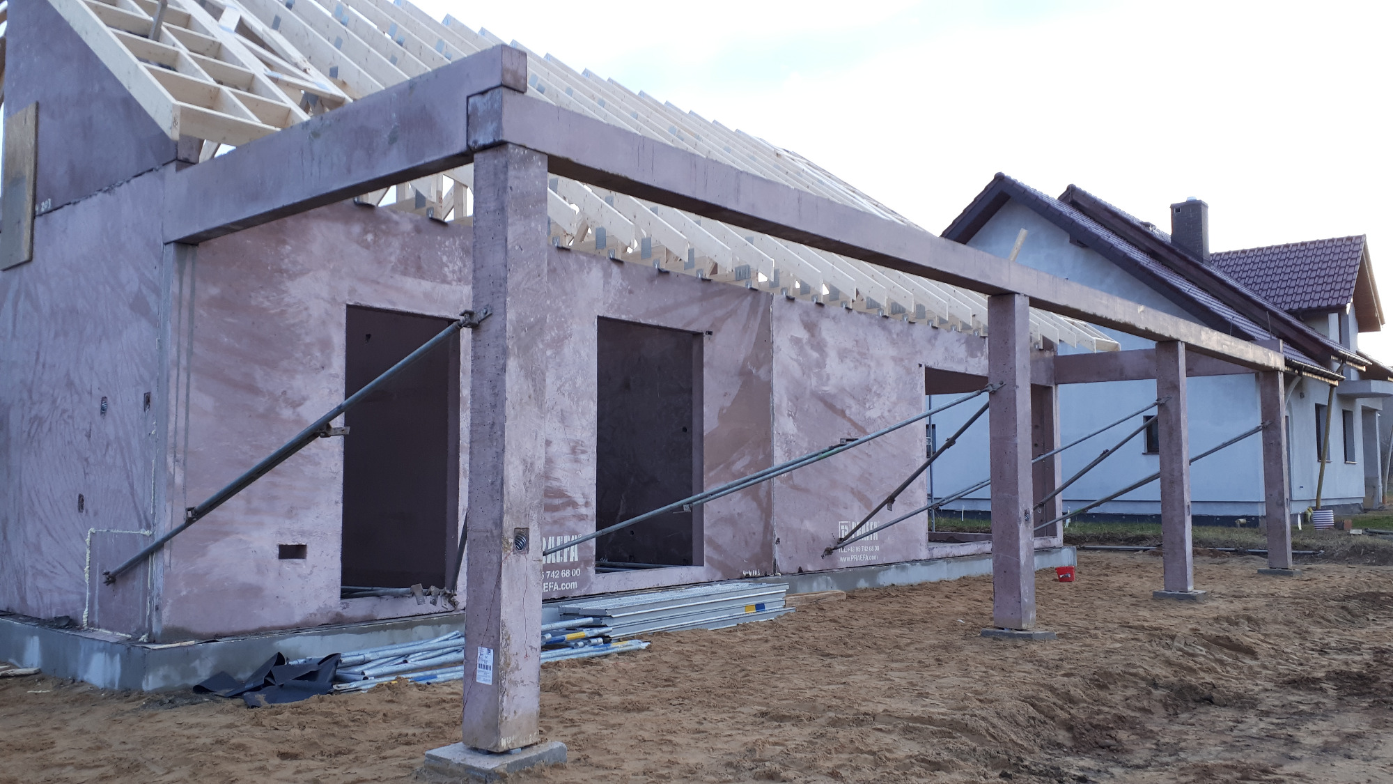 Surowy stan domu murowanego z widocznym szkieletem dachu, wspornikami i ścianami otynkowanymi na różowo, na tle sąsiedniego domu.