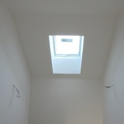 Gfk Design Grzegorz Kowalski - Białe, świeżo pomalowane poddasze z oknem dachowym. Widoczne wystające kable i otwór na gniazdko, sugerujące prace wykończeniowe.